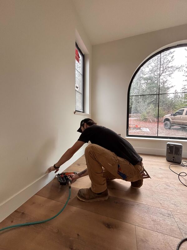 A man kneels on a wooden floor installing white baseboards with a nail gun in a room with large windows.